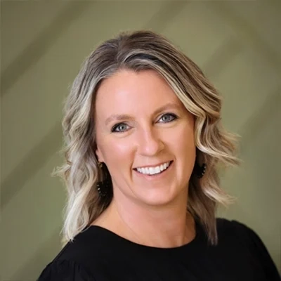 Headshot of a smiling woman with wavy blonde hair, blue eyes, and a black top, against a blurred olive green background.
