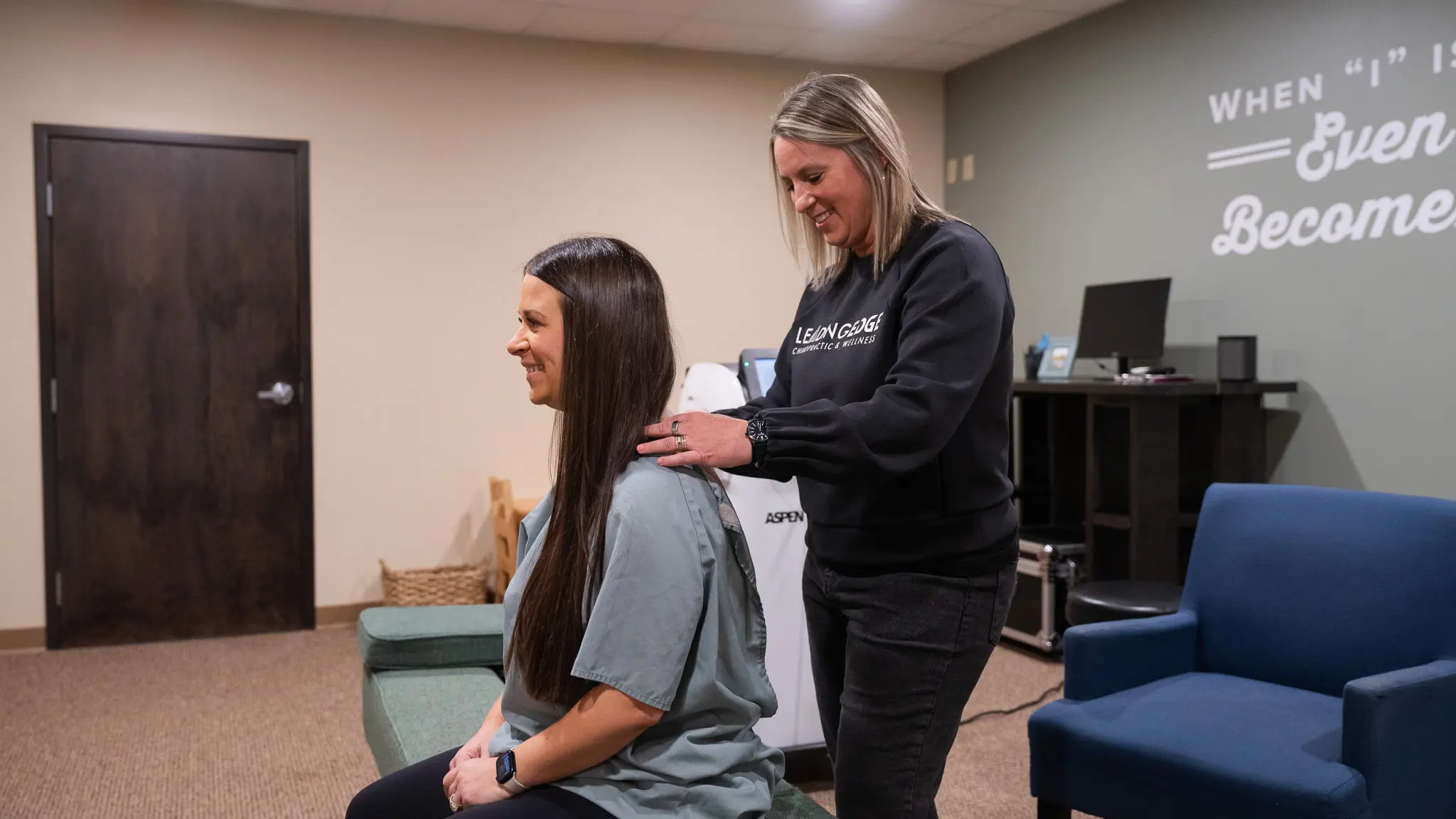 A chiropractor in a black sweatshirt gently places her hands on the shoulders of a smiling patient with long dark hair, who is seated on a green bench in a modern clinic room.