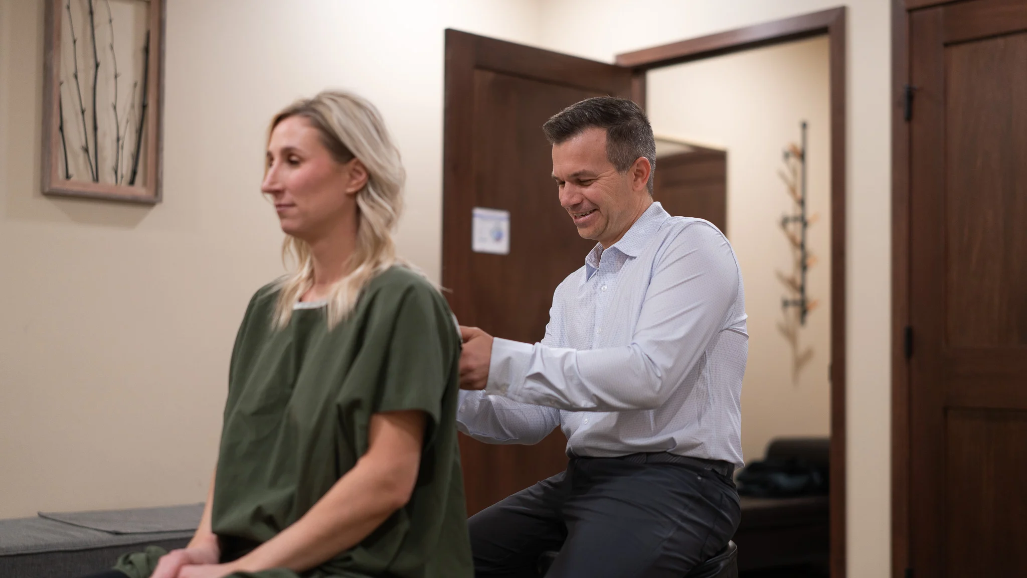 A male chiropractor or therapist smiling while examining the upper back of a female patient in a green gown.