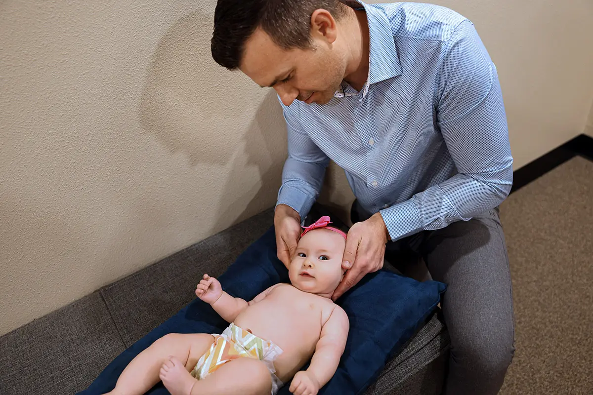 A man in a blue shirt gently holds the head of a baby lying on a blue pillow. The baby wears a pink bow and a patterned diaper, looking at the camera.