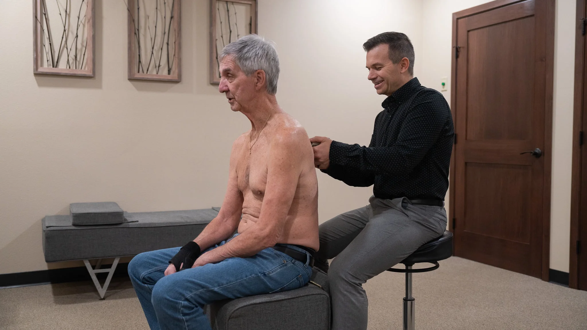 An older man with gray hair sits shirtless, receiving back therapy from a male practitioner who is smiling and working on his upper back.