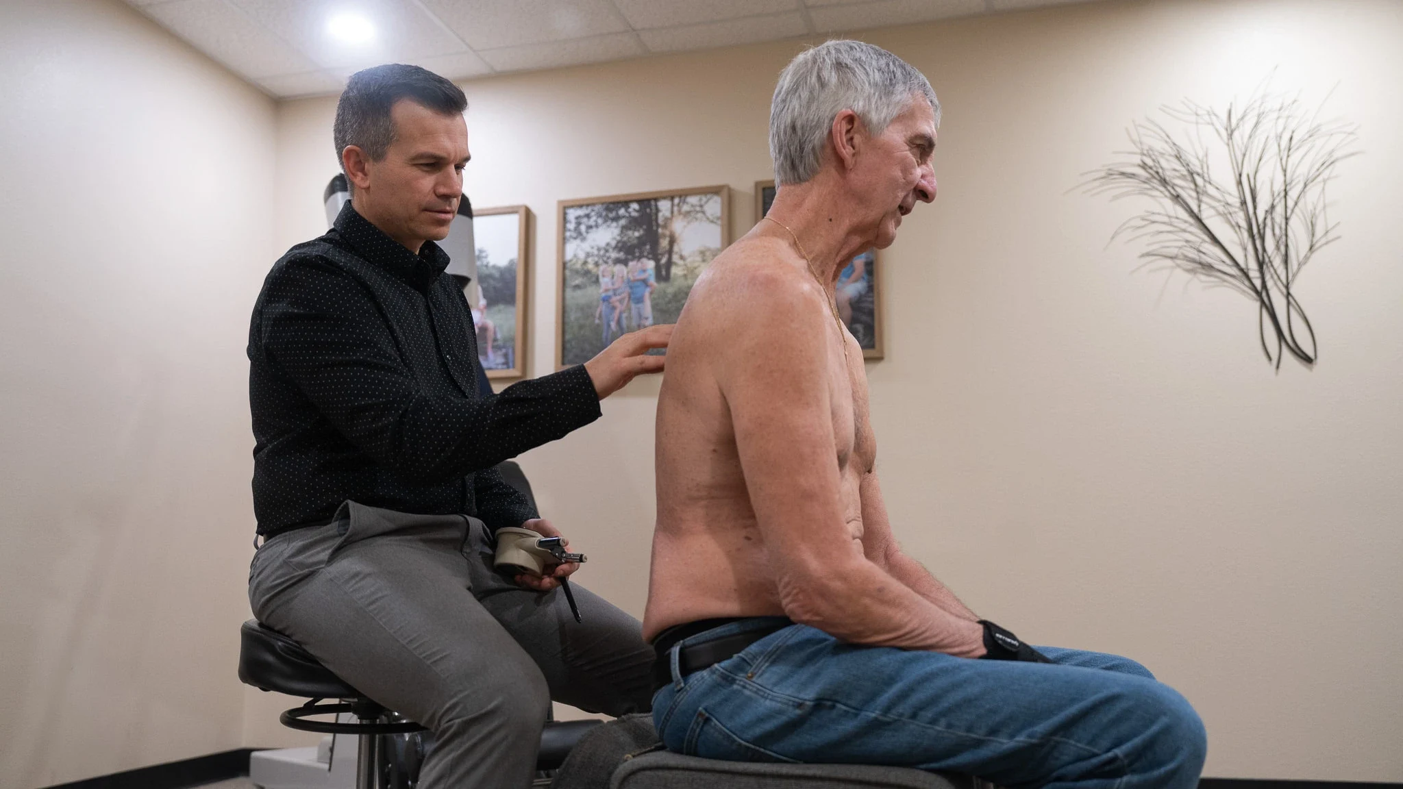 A male chiropractor in a black shirt examines the bare back of an older male patient in blue jeans, who is seated in profile. The chiropractor holds a small tool in his other hand. Framed pictures and a wall sculpture are in the background.