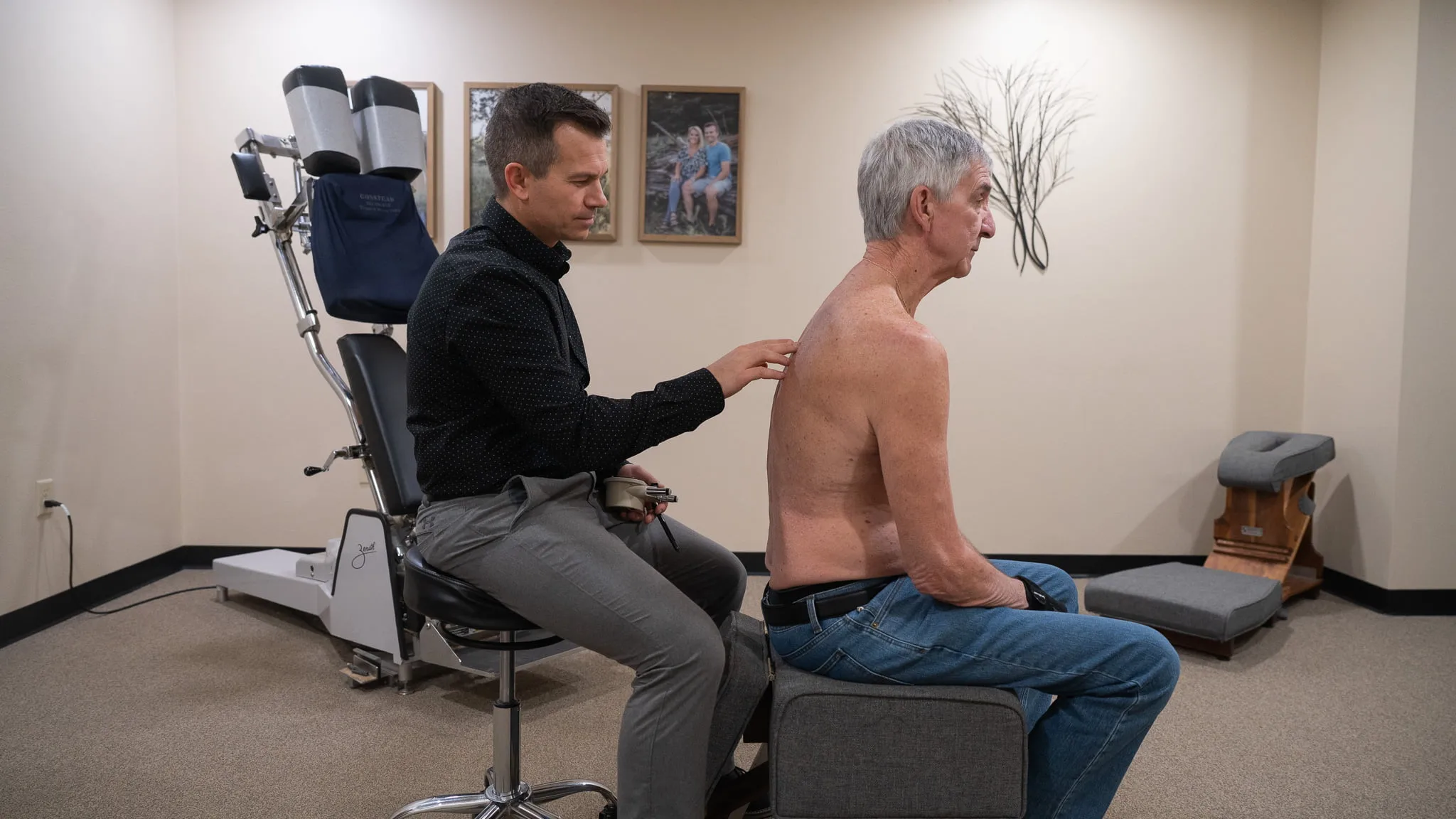 A male chiropractor examines the bare upper back of an older male patient sitting on a stool in a chiropractic office.