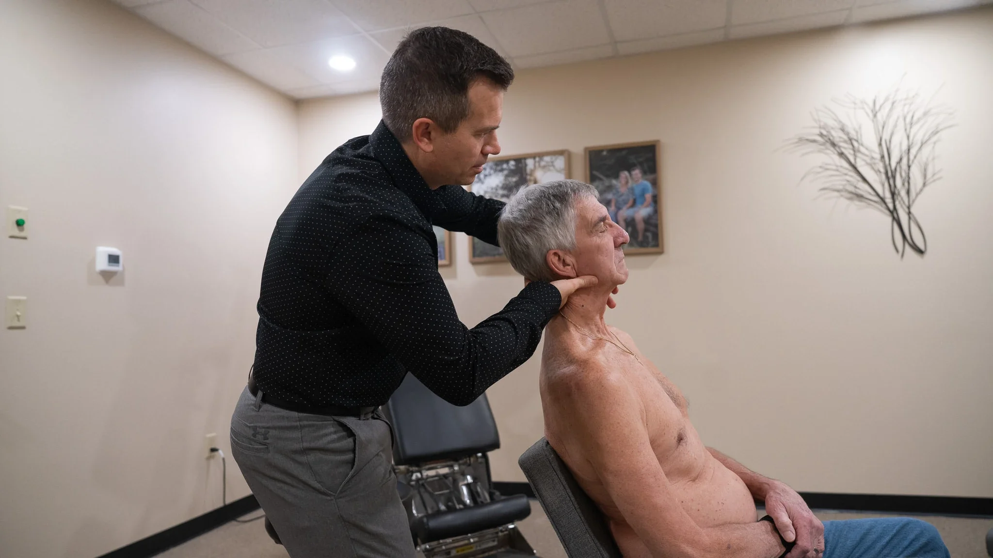 A chiropractor in a black polka-dot shirt performs a neck adjustment on an older, shirtless male patient with grey hair, who is seated in a chair.