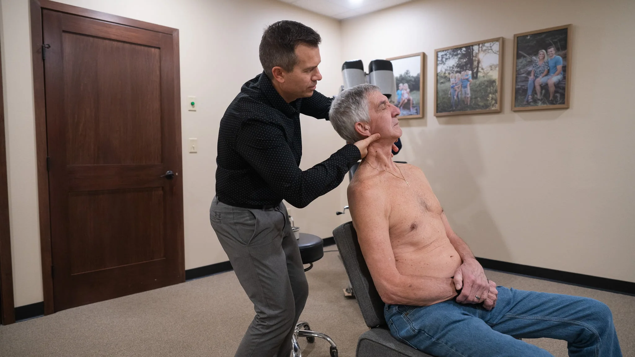 A chiropractor, wearing a black polka dot shirt, adjusts the neck of an older male patient who is shirtless and seated in a gray chair. The room has beige walls and framed photos.