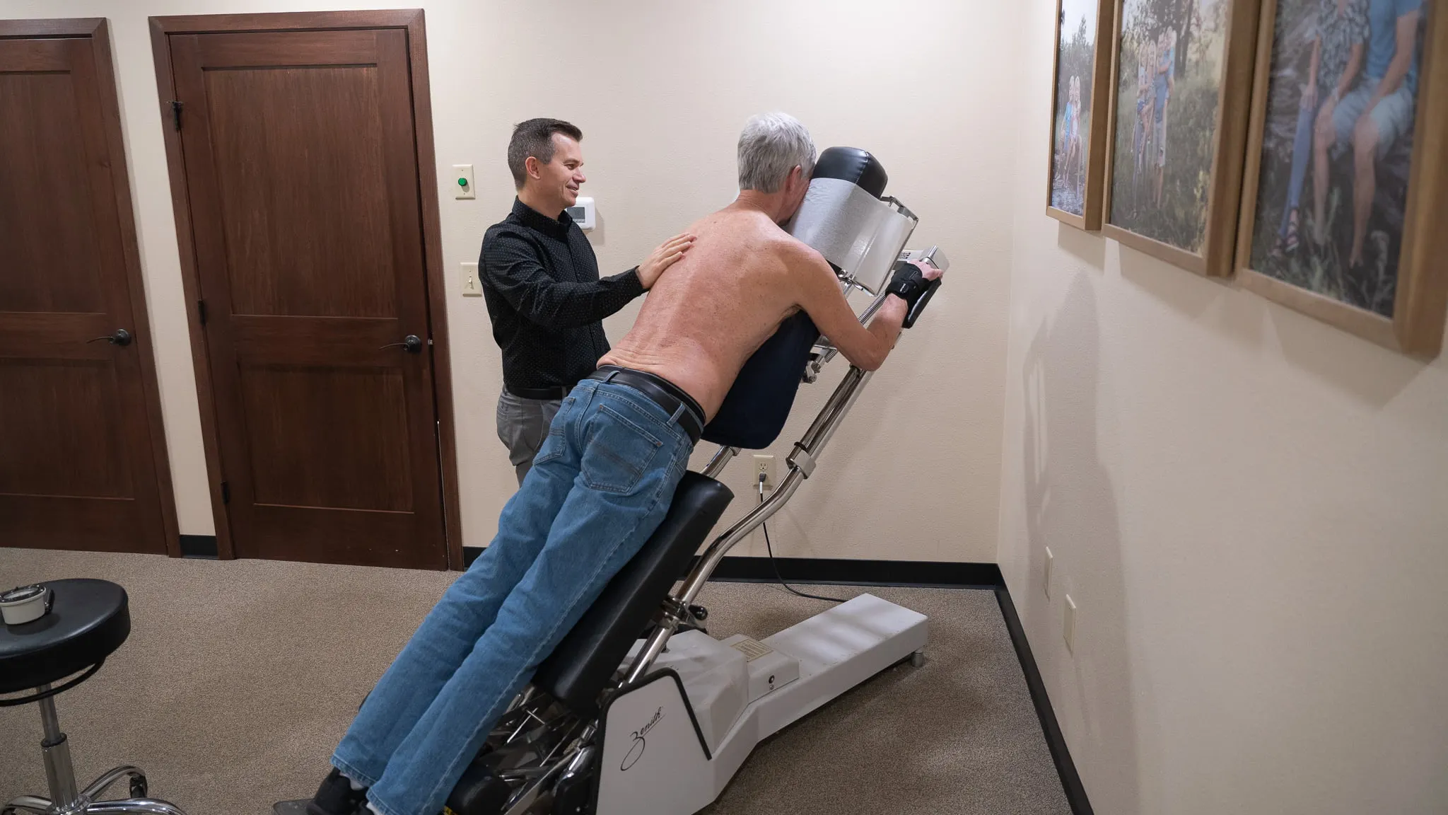 A male practitioner with a smiling face gently touches the back of an older male patient who is lying face down on a chiropractic treatment table in a clinic room.