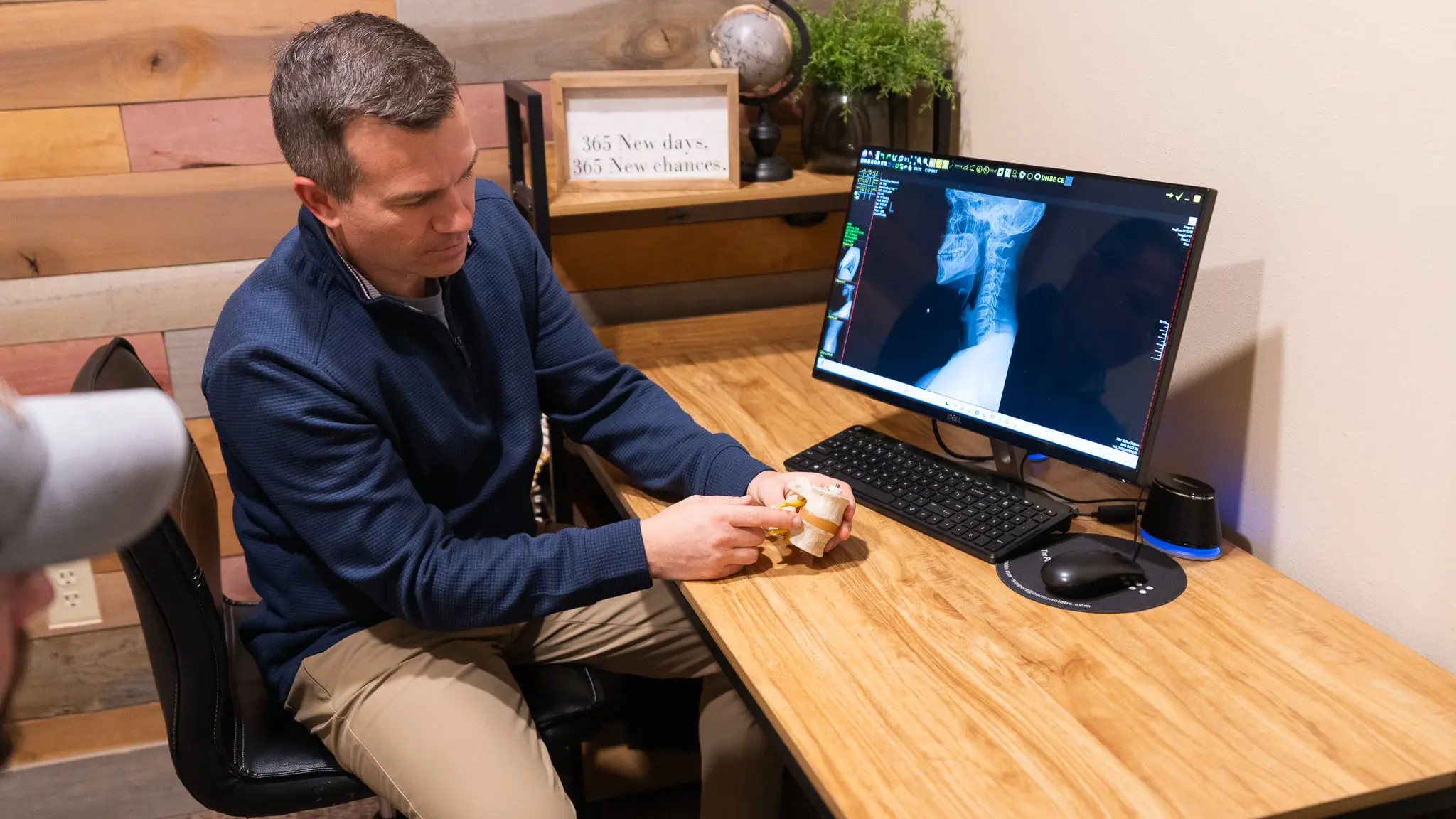 A man in a blue shirt holds a spinal model while looking at a neck X-ray displayed on a computer monitor, explaining medical information.