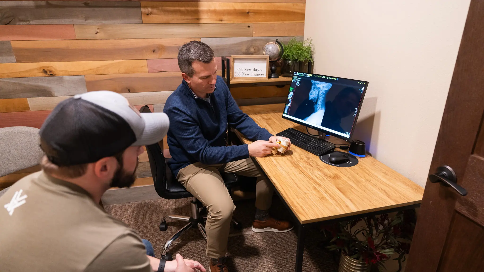 A practitioner explains a spine model to a patient, with a neck X-ray visible on a computer monitor on the desk in a modern office.
