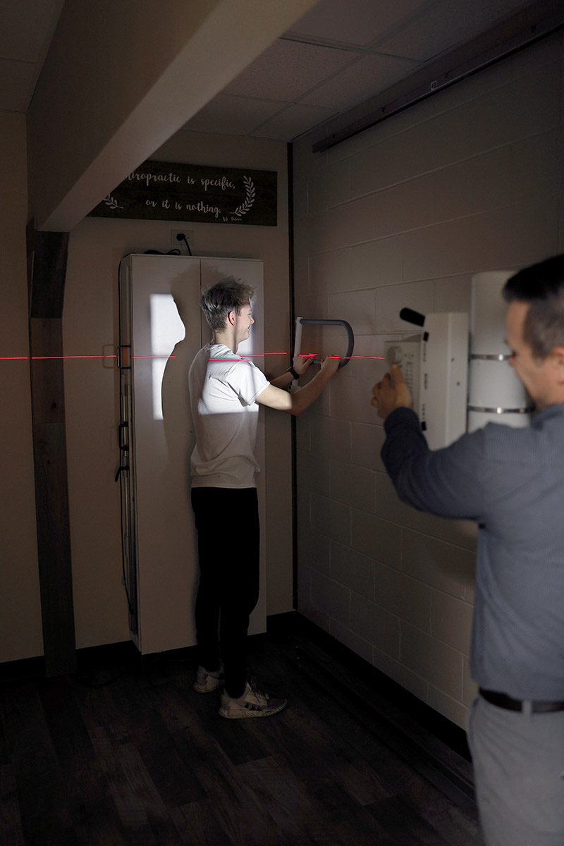 A young man stands against a wall with a red laser line across his upper back, undergoing a posture assessment, while a practitioner operates equipment. A sign above reads 'Chiropractic is specific, or it is nothing.'