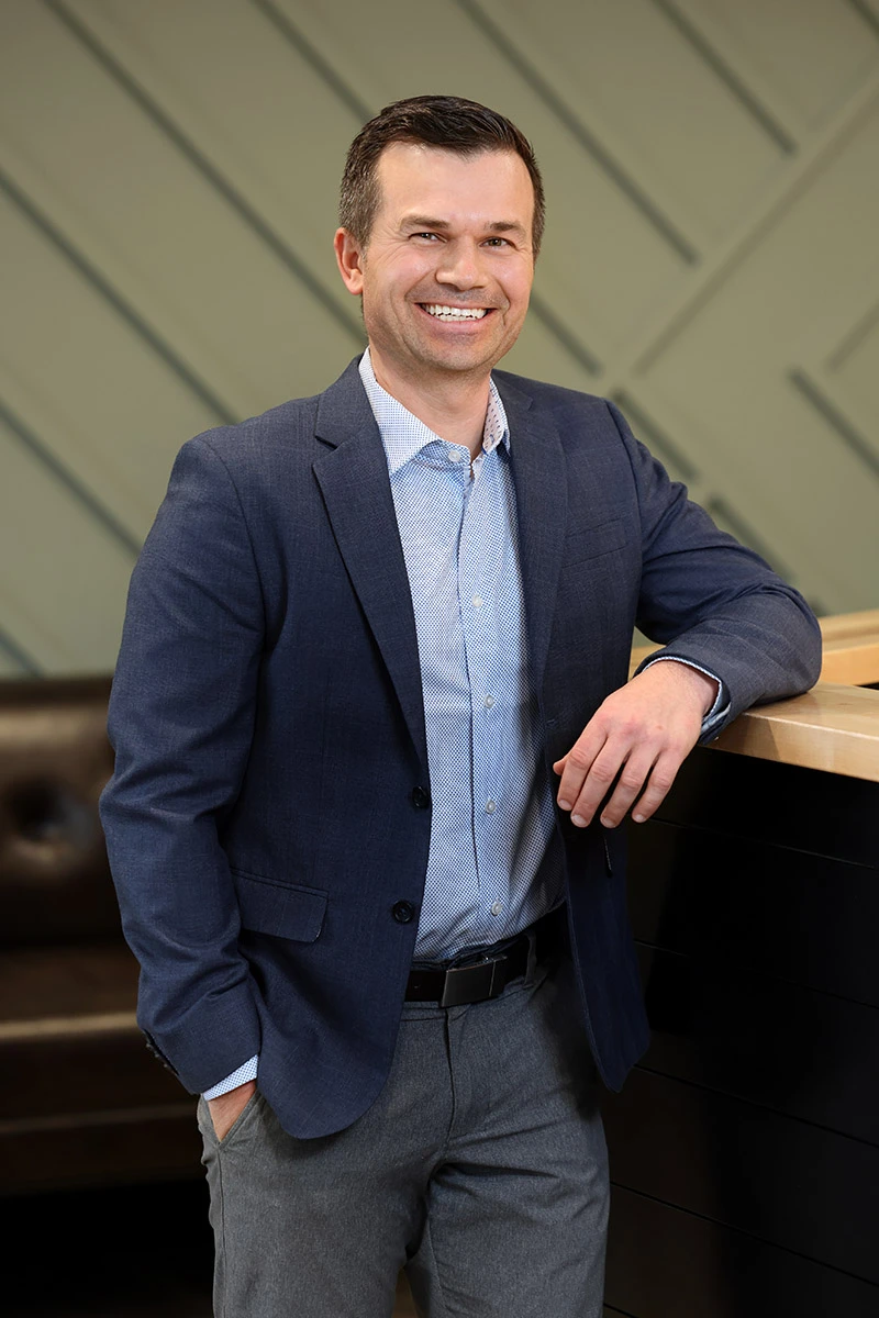 A smiling man in a blue blazer and light blue shirt, standing with one hand in his pocket and the other resting on a counter.
