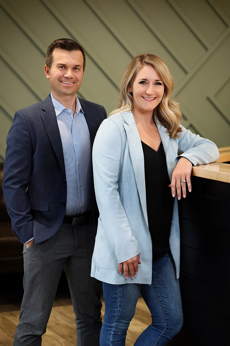 A smiling man in a blue blazer and a woman in a light blue blazer and jeans stand in a modern office setting with a geometric green wall.