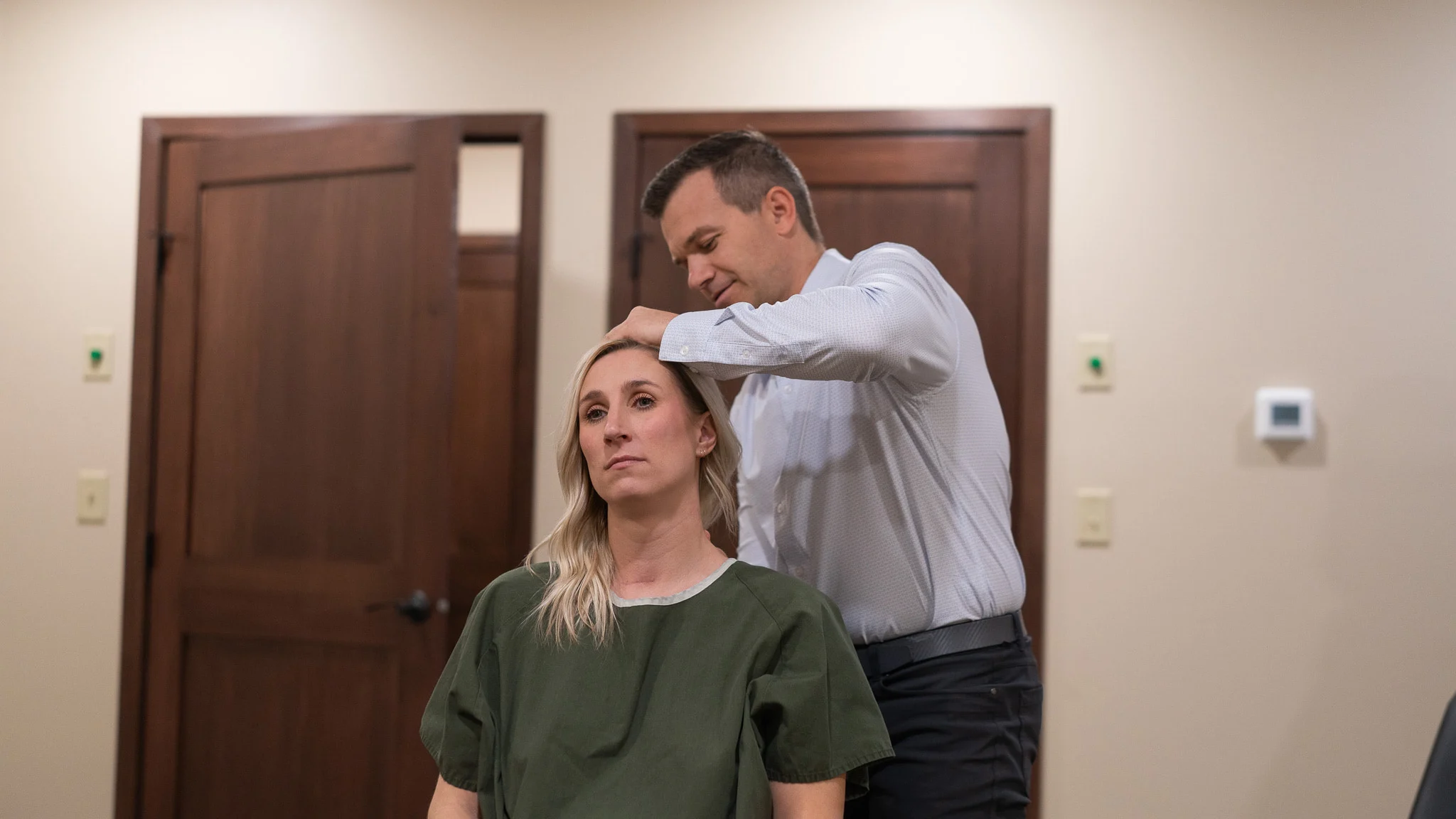 A man in a light blue shirt places his hands on the head of a blonde woman in a green top, performing an adjustment. They are in a room with wooden doors.