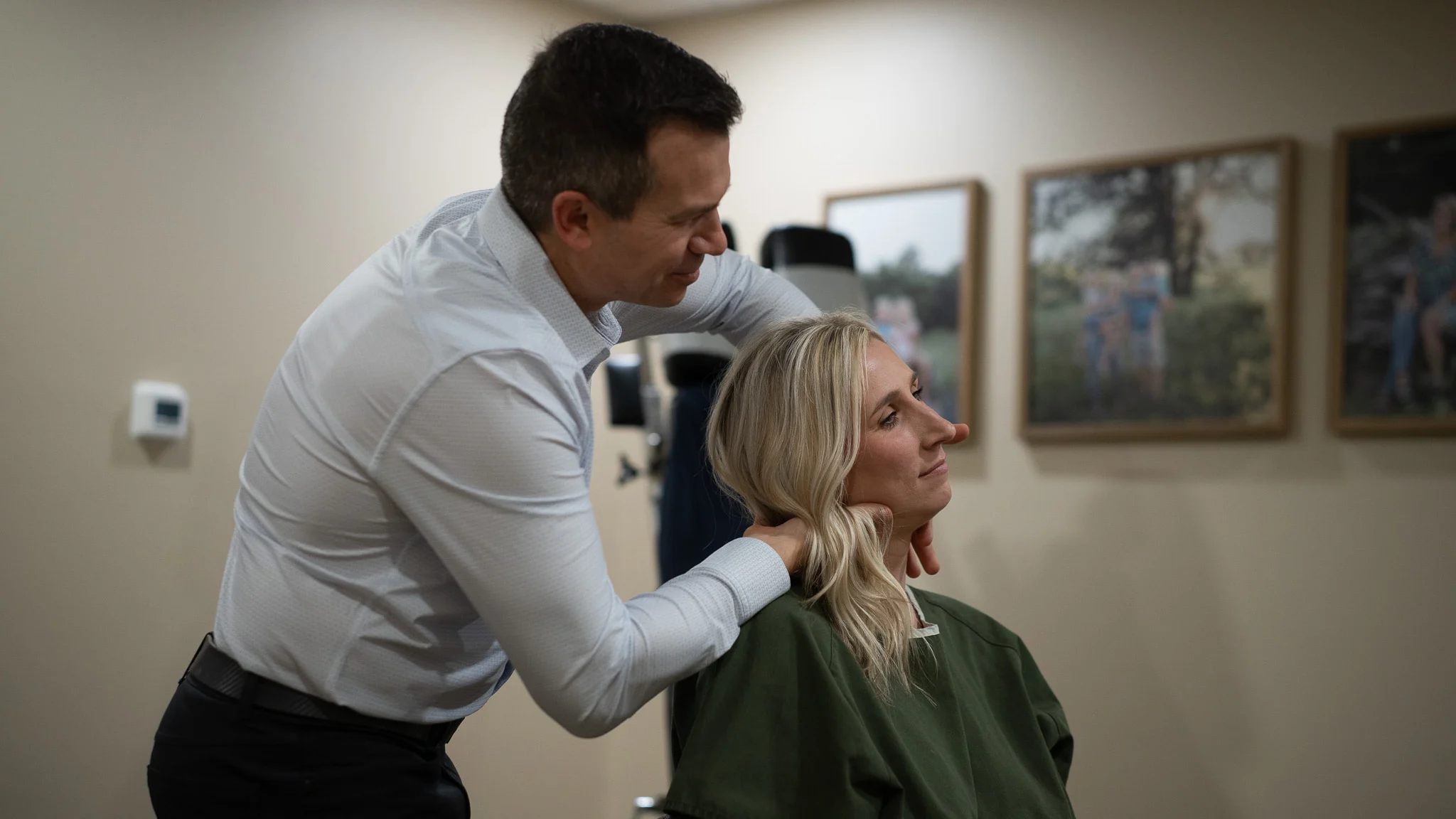 A male chiropractor adjusts a female patient's neck, with his hands on her shoulders, in a clinic room with framed pictures on the wall.