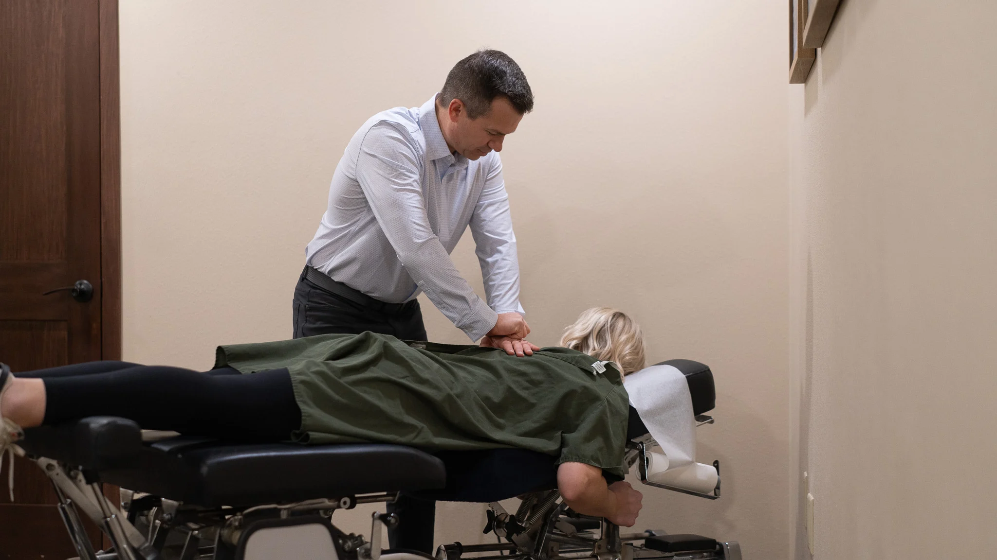 A male chiropractor adjusts the back of a female patient lying face down on a black chiropractic table in a treatment room.