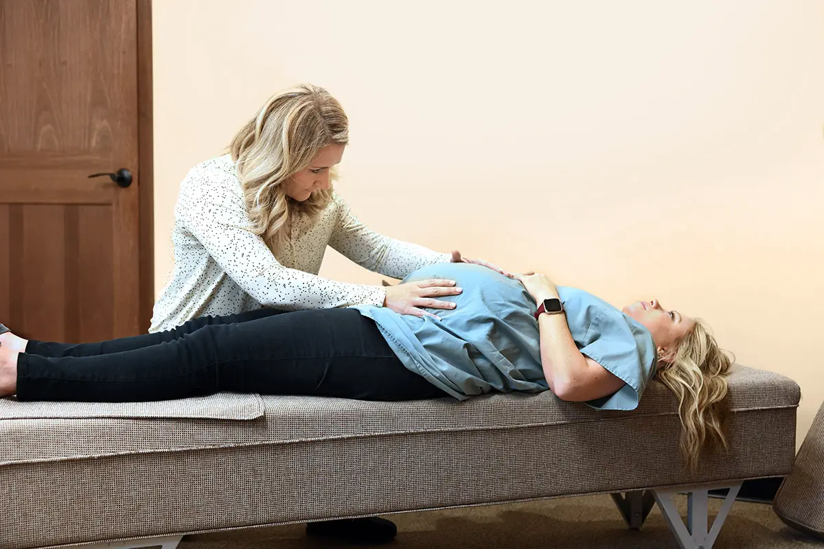 A practitioner with blonde hair gently touches the pregnant belly of a woman lying on a treatment table.