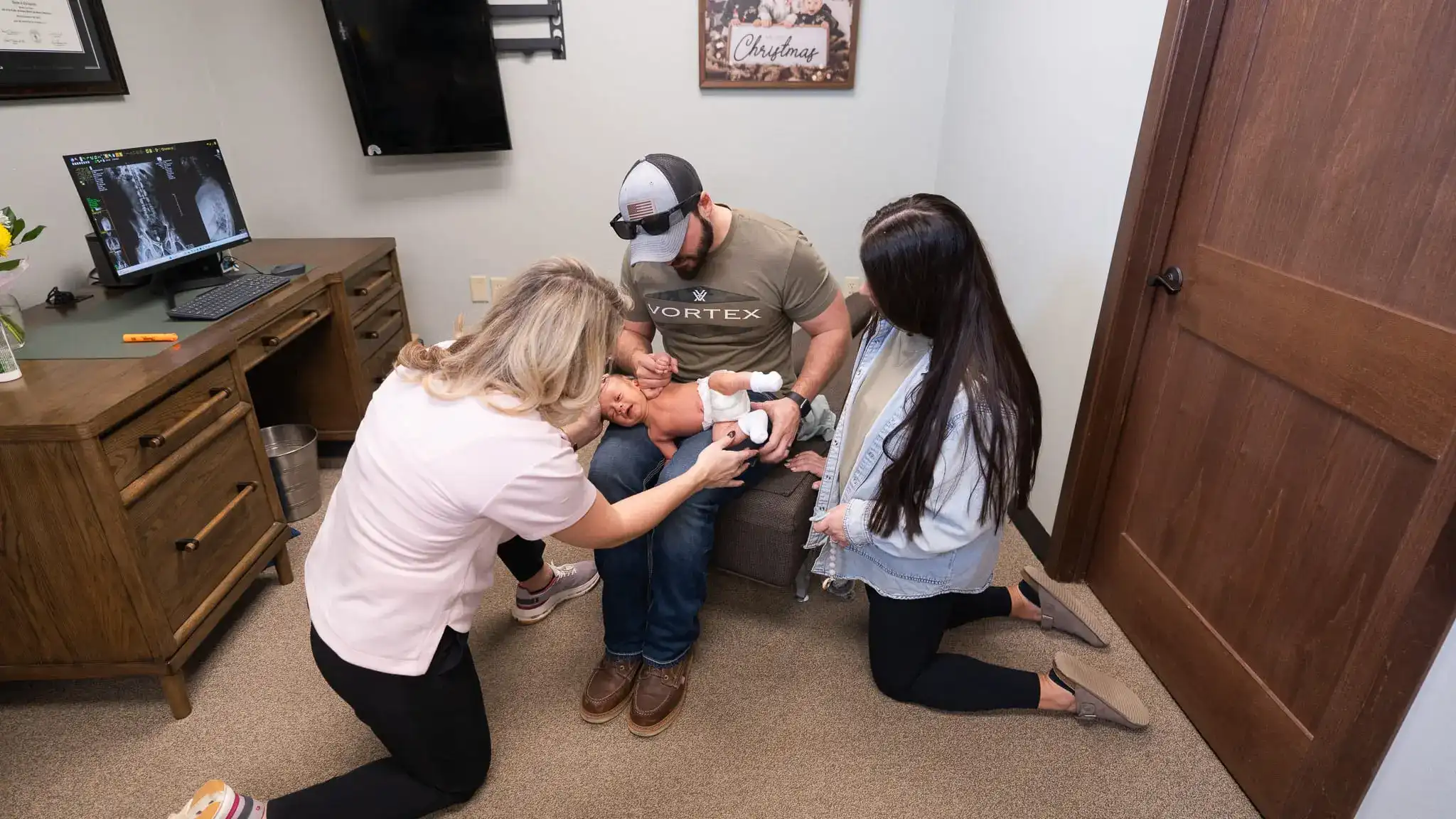 A chiropractor kneels to gently examine a newborn baby held by its father, while the mother watches. A computer screen with spinal X-rays is visible in the background.