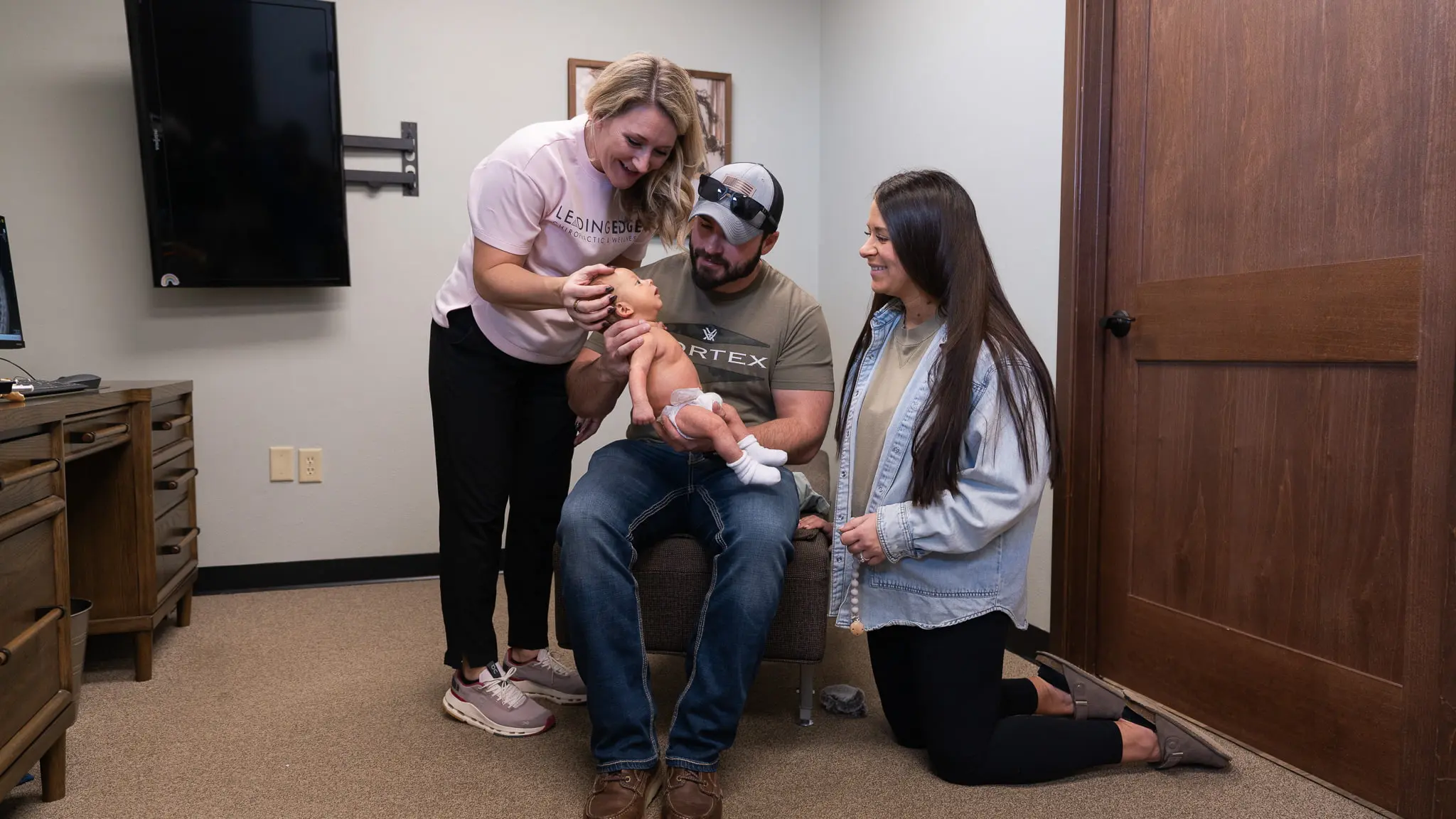 A female chiropractor gently adjusts a baby held by a seated man, while a woman kneels beside them, all smiling at the infant in a clinic setting.
