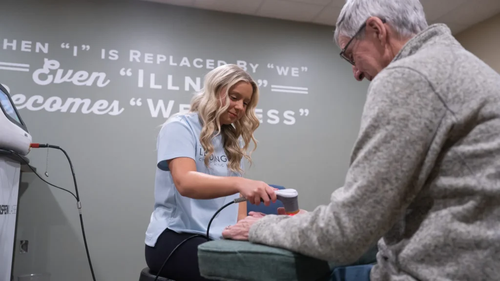A female therapist uses a red light laser device to treat an older man's hand in a clinic, with a motivational quote about wellness on the wall.