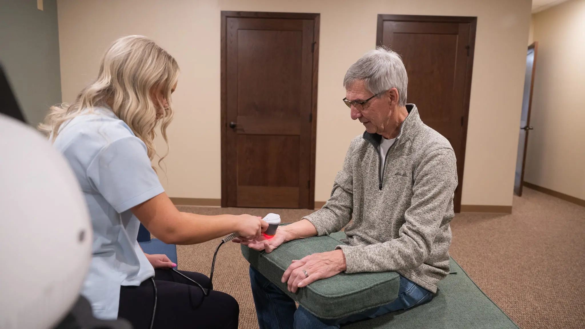 A therapist applies a red light therapy device to an older man's wrist and hand, which rests on a green cushion, in a clinic setting.