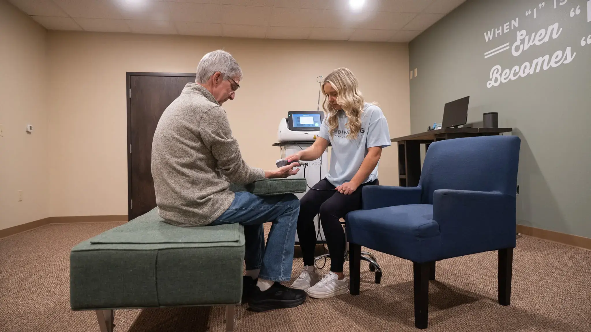 An older man sitting on a bench receives laser therapy on his wrist from a female therapist in a modern clinic room.