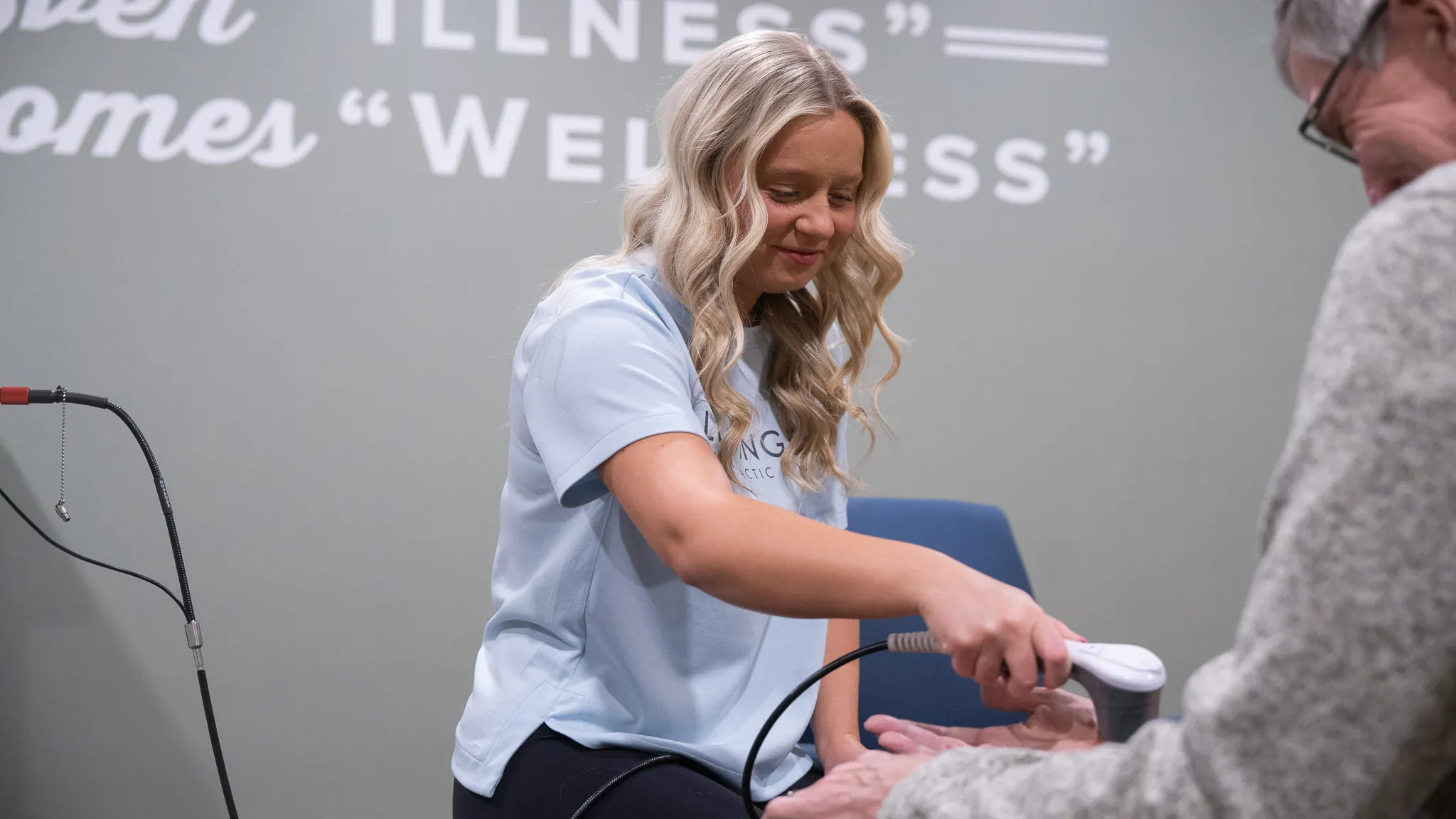 A blonde woman in a light blue chiropractic shirt uses a handheld device to treat an older person's hand. White text on the grey wall behind them reads 'ILLNESS' and 'WELLNESS'.