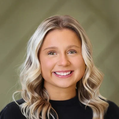 Headshot of a young woman with long blonde wavy hair smiling at the camera against a soft green background.