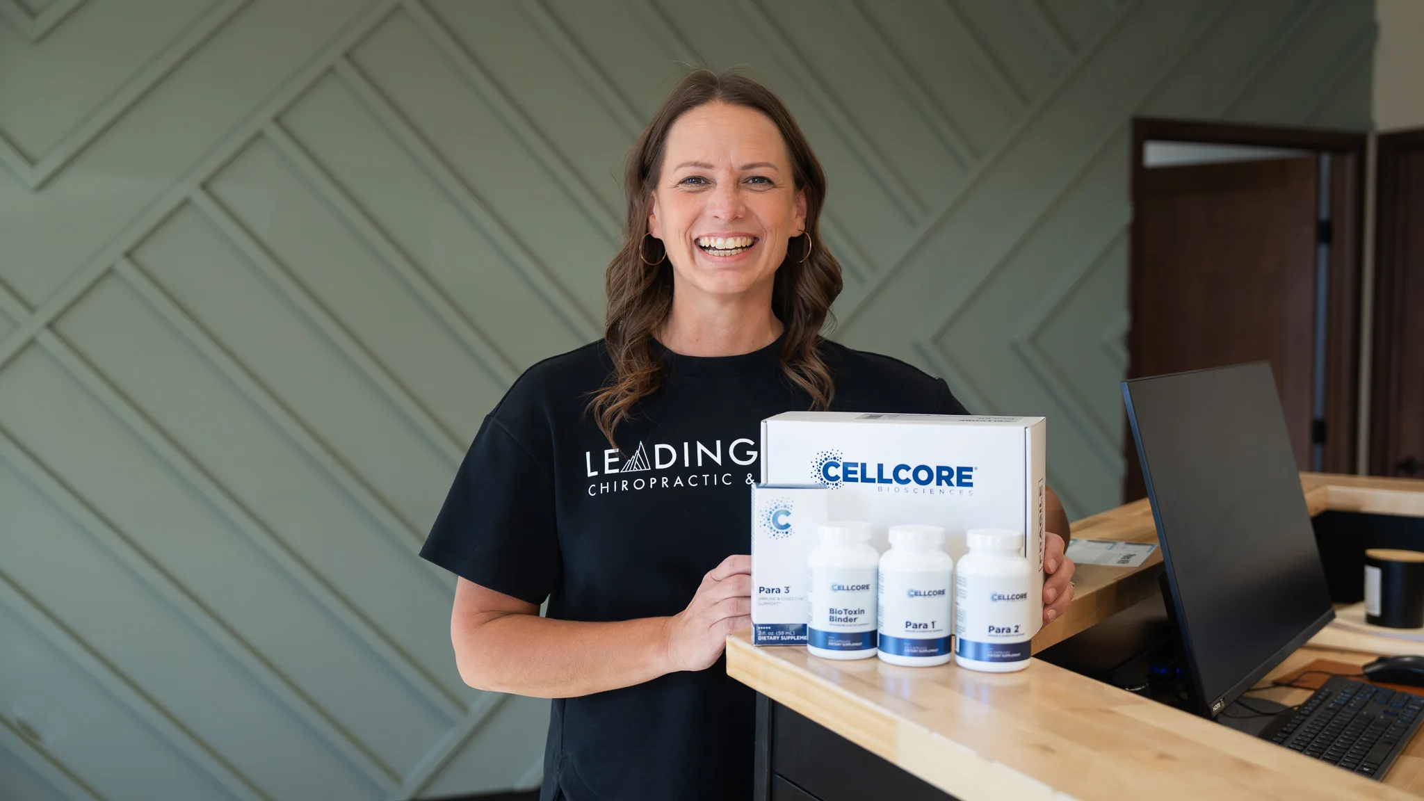 A smiling woman in a black 'Leading Chiropractic' t-shirt stands behind a desk, holding a box and bottles of CellCore Biosciences supplements, including BioToxin Binder and Para products.