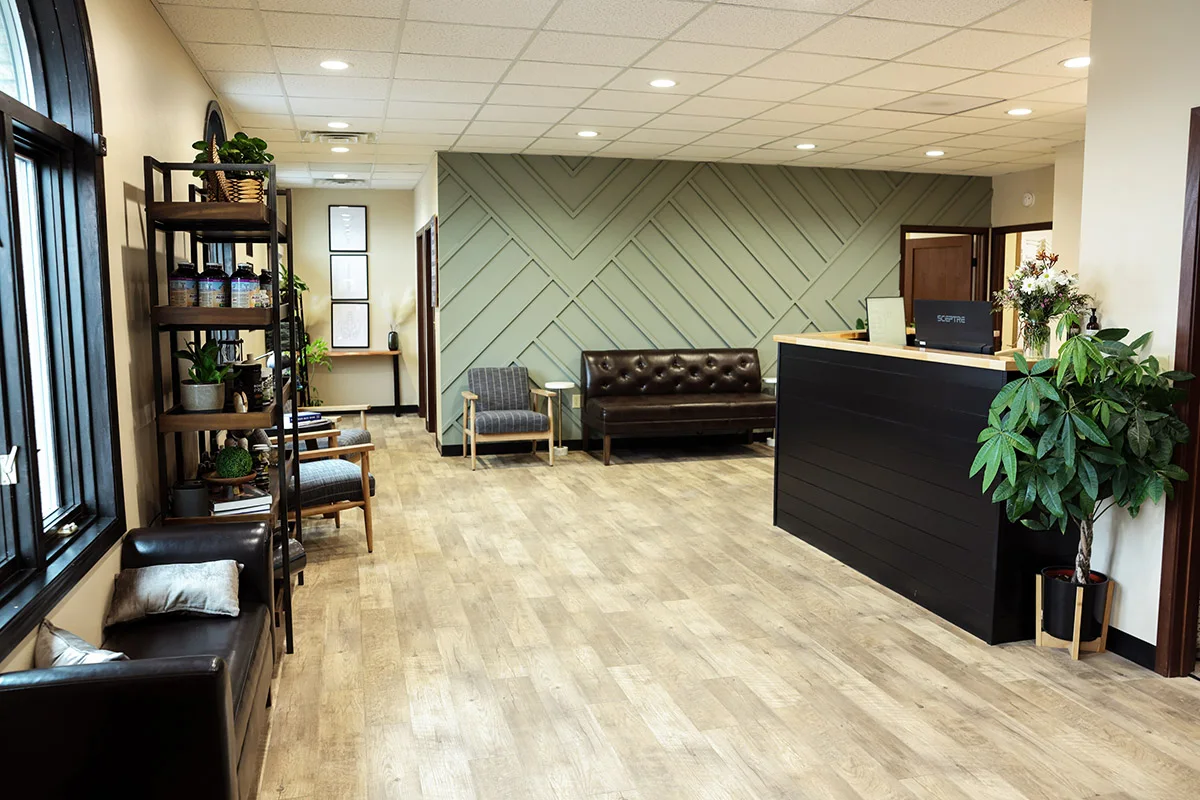 A modern and inviting reception area with a sage green geometric accent wall, comfortable brown leather and striped fabric seating, a black reception desk, and several potted plants.