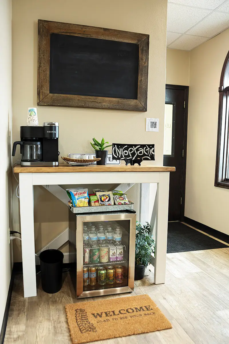 A chiropractic office refreshment station featuring a Keurig coffee maker, a snack tray, a mini-fridge stocked with drinks, and a welcome mat that reads 'Glad to see your back'.