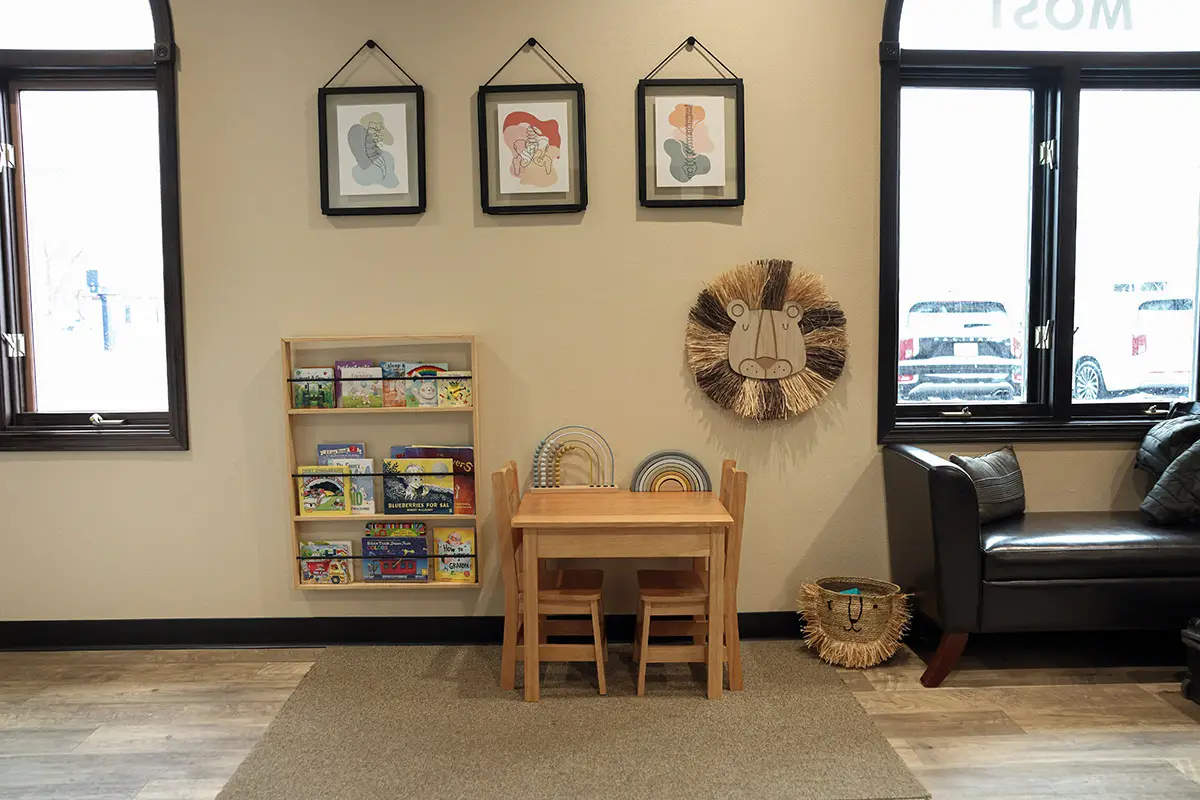 A bright children's waiting area with a small wooden table and chairs, a bookshelf filled with books, and lion-themed decor, including a wall hanging and a basket. Two large windows let in natural light.