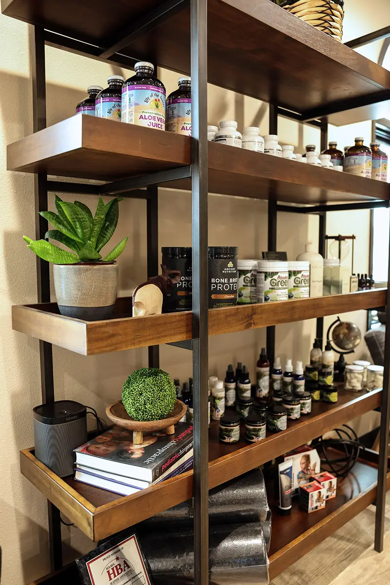 A multi-tiered dark wood and black metal shelf unit filled with various wellness products, including aloe vera juice, protein powders, a snake plant, books, a Sonos speaker, and small bottles of oils.