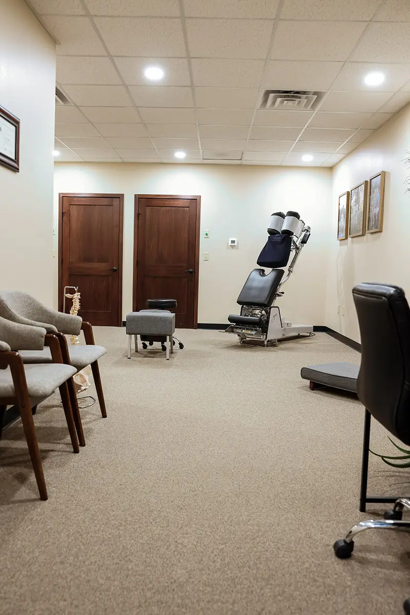 A clean chiropractic treatment room with an adjustment table, two grey chairs, and two wooden doors.