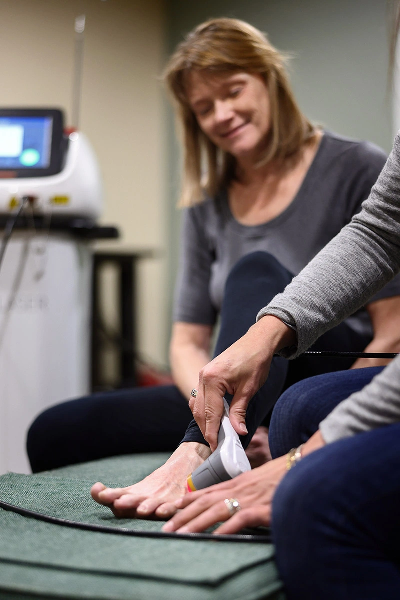 Close-up of a bare foot receiving laser therapy from a handheld device, with a smiling practitioner blurred in the background.