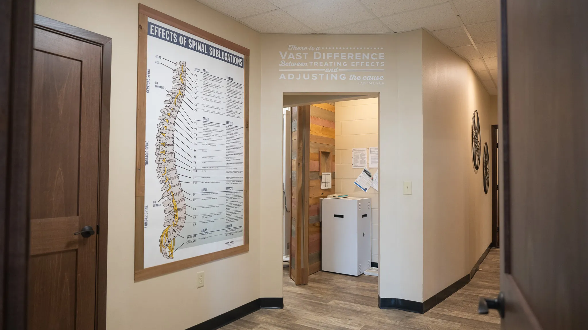 A chiropractic clinic hallway with a spinal subluxation chart on the left wall and a quote by D.D. Palmer on the right wall: 'There is a vast difference between treating effects and adjusting the cause.'