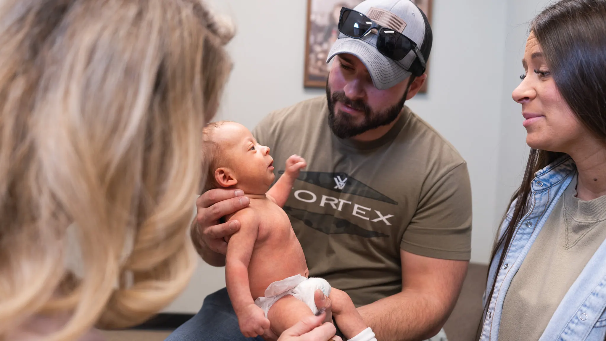 A man in a cap and t-shirt gently holds a small baby in a diaper, looking at it with affection. A woman with long dark hair smiles beside them, and another person with blonde hair is partially visible on the left.