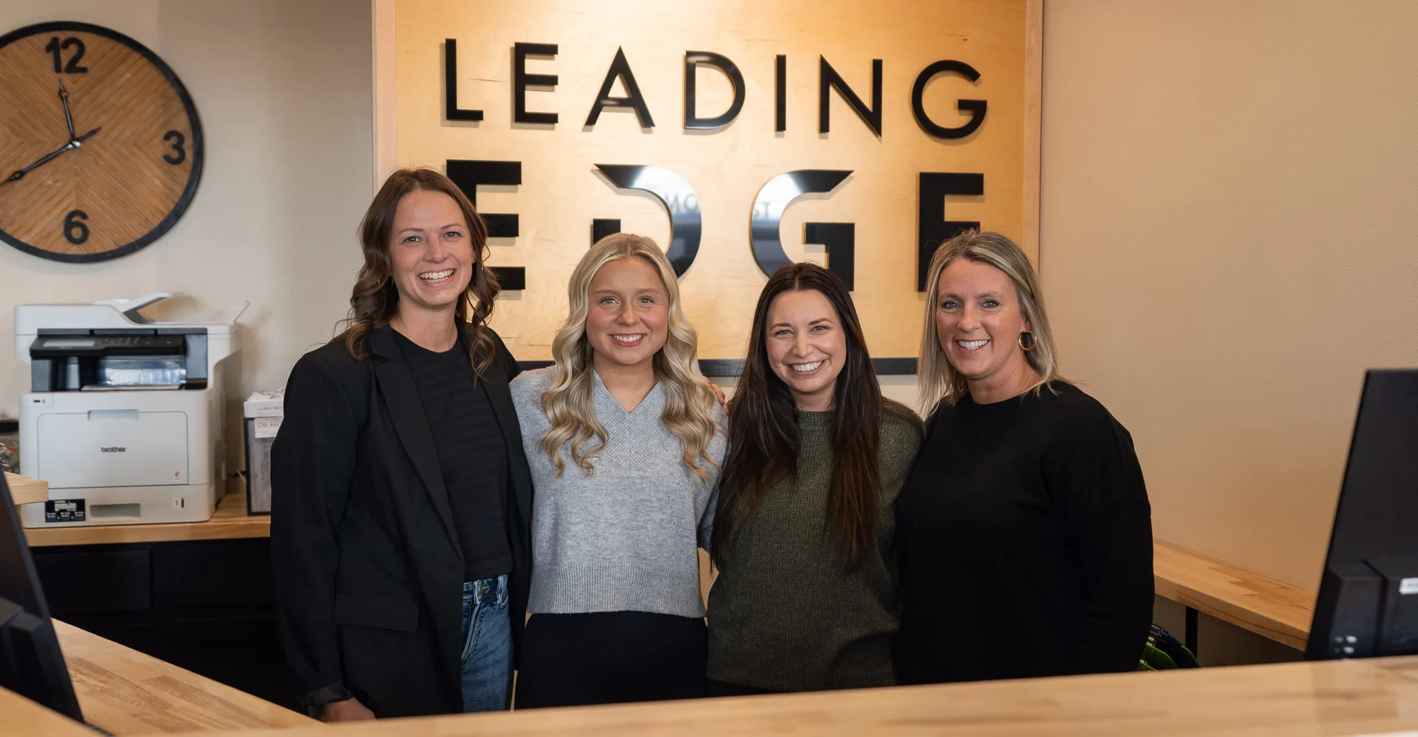 Four smiling women stand behind a wooden counter in an office. Behind them is a large sign that reads 'LEADING EDGE' and a wooden clock.