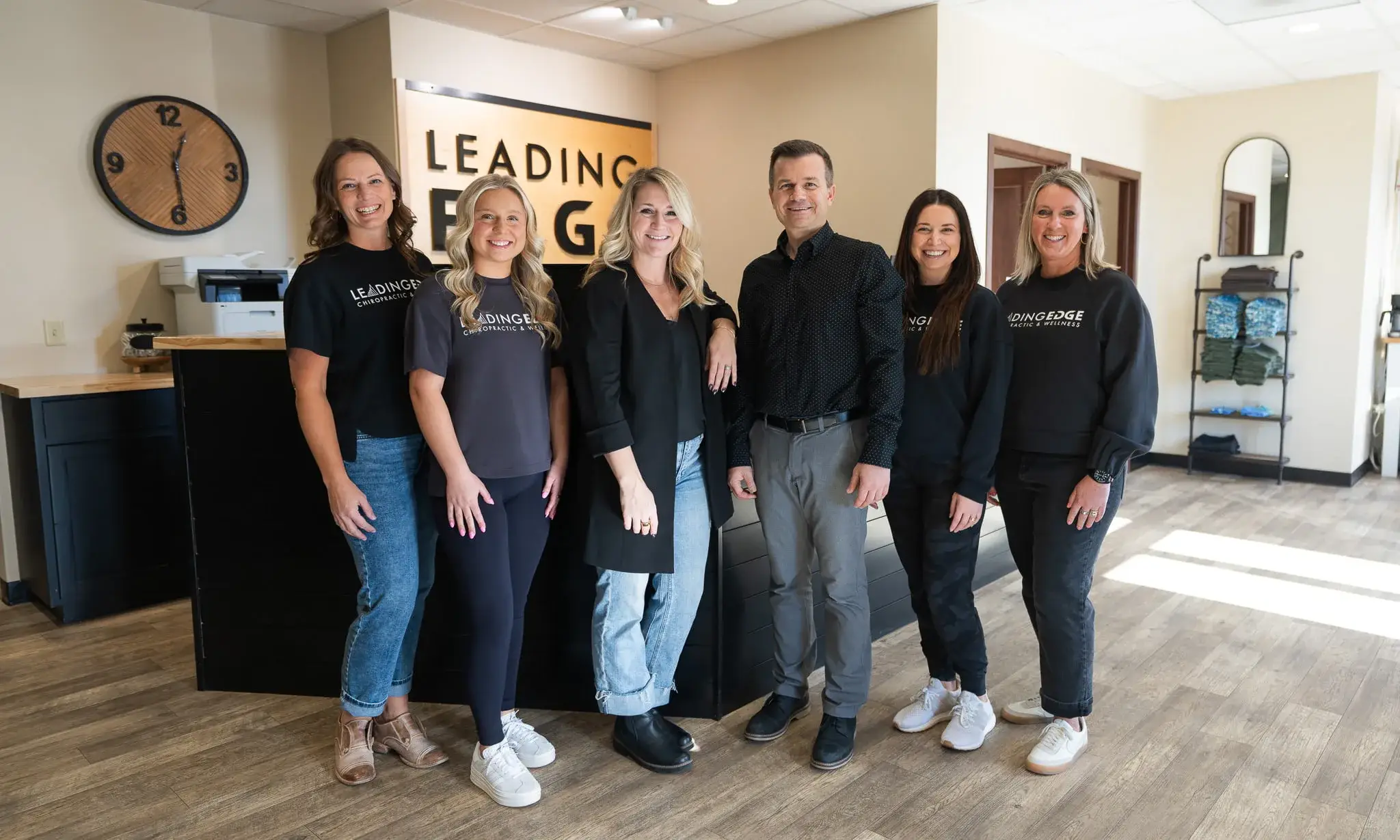 Six smiling team members, five women and one man, stand in the modern reception area of LeadingEdge Chiropractic & Wellness. They wear branded shirts, and a large 'LeadingEdge' sign is visible behind them.