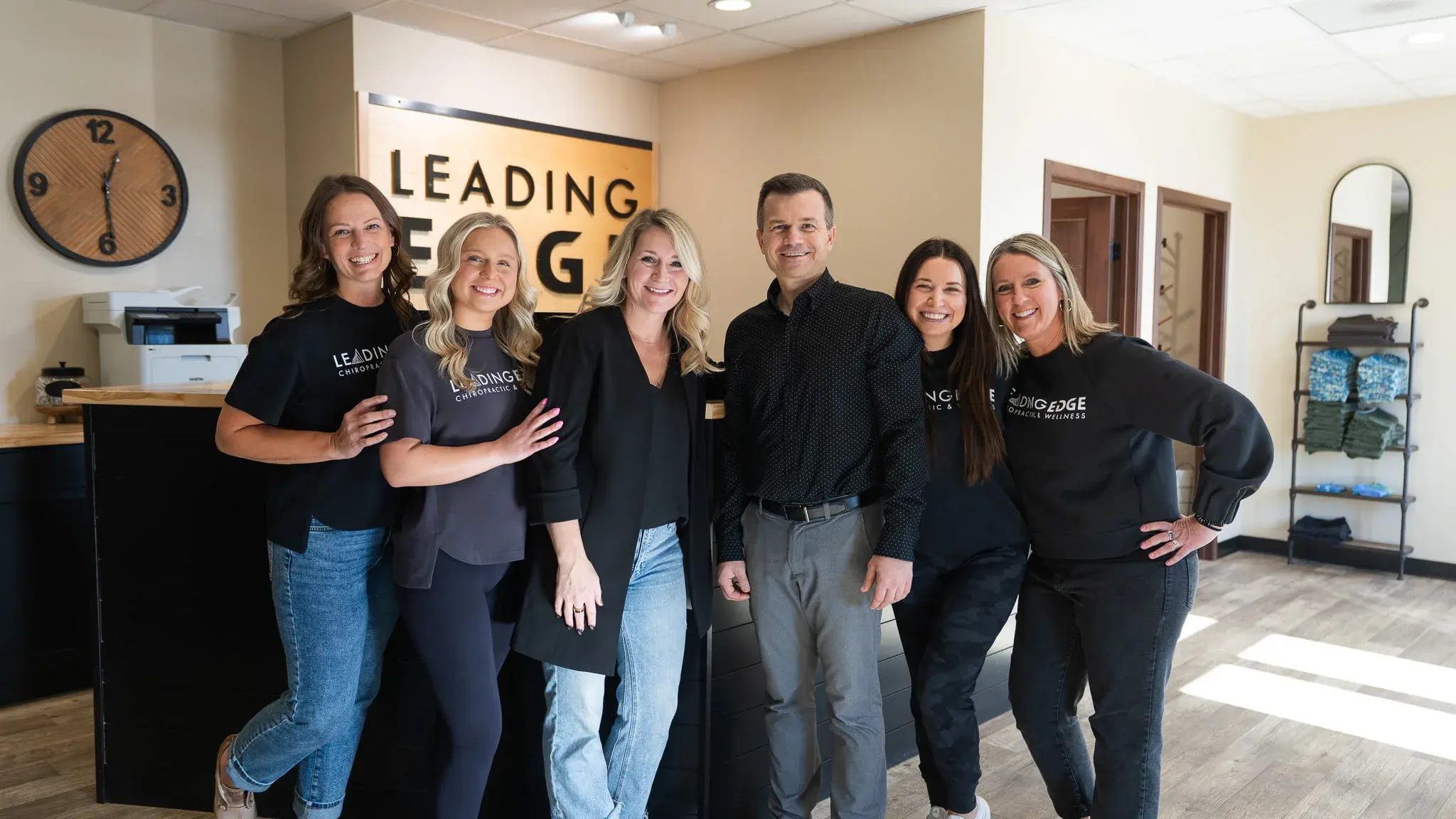 Group photo of six smiling team members, five women and one man, standing in the modern reception area of Leading Edge Chiropractic & Wellness clinic.
