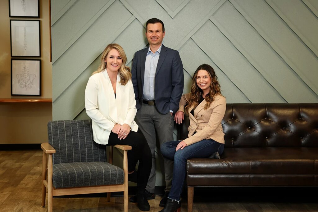 Three smiling professionals, two women and one man, in a modern office setting with a green paneled wall and anatomical artwork.