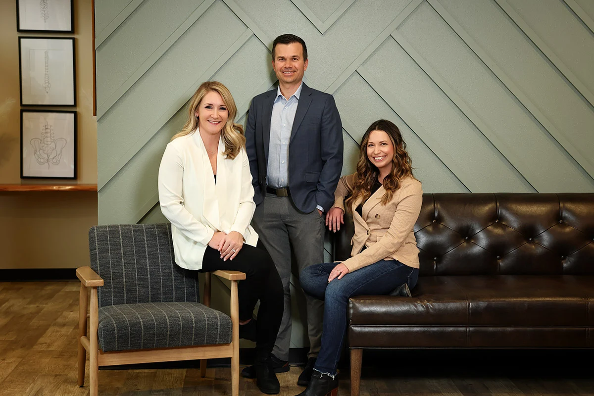 Three smiling professionals, two women and one man, in a modern office setting with a green paneled wall and anatomical artwork.