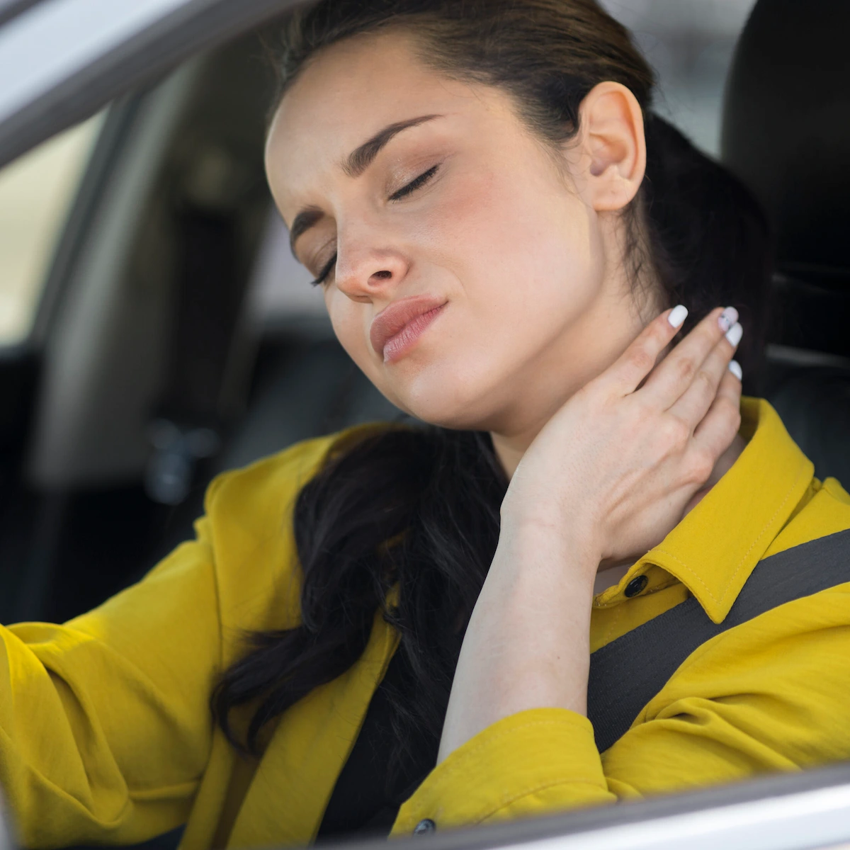A woman in a yellow shirt sits in a car with her eyes closed, holding her hand to her neck, indicating pain or discomfort.