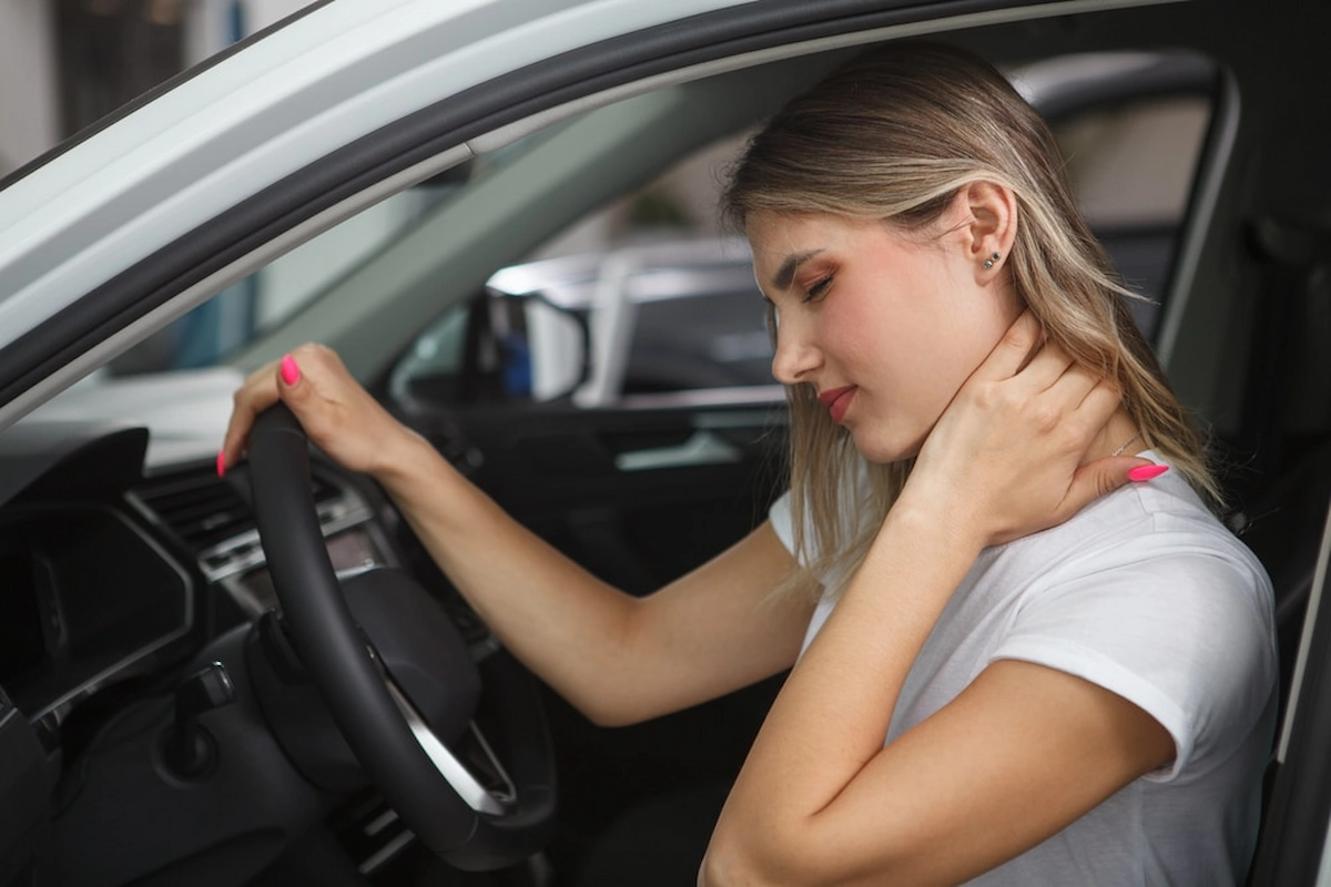 A woman in a white t-shirt sits in a car, holding her neck with her right hand and the steering wheel with her left, looking down with an expression of discomfort or pain.