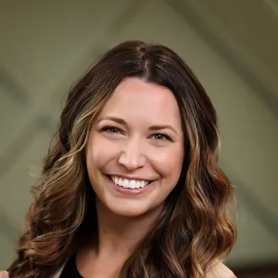 Headshot of a smiling woman with long, wavy brown hair and light eyes, wearing a black top and a light blazer.