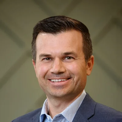 Headshot of a smiling man with short dark hair, wearing a blue blazer and patterned shirt.