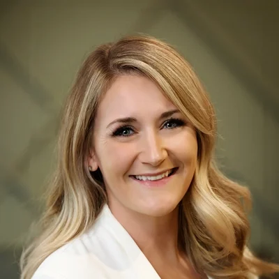Headshot of a smiling woman with long, wavy blonde hair and blue eyes, wearing a white blazer.