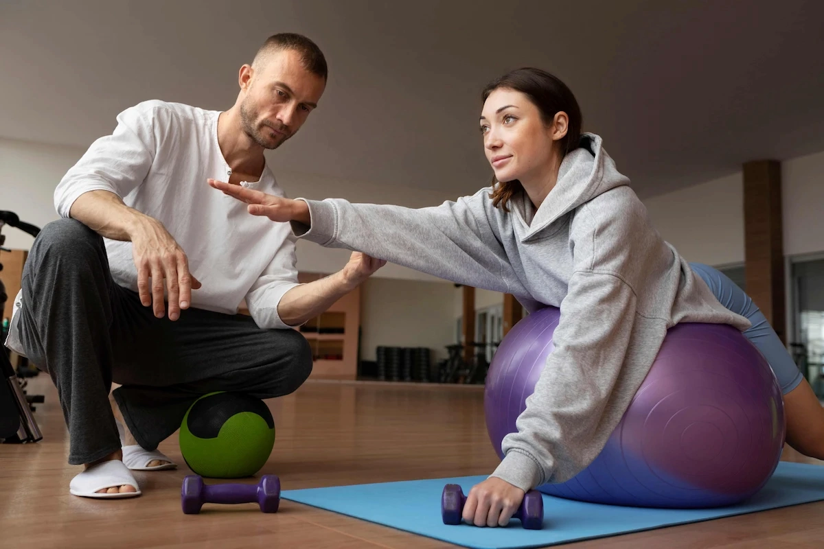 A male trainer in a white shirt guides a woman on a purple stability ball, who is holding a dumbbell and extending her arm during an exercise in a gym.