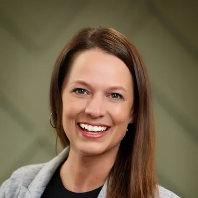 Headshot of a smiling woman with long brown hair, wearing a gray blazer, against a blurred green background.