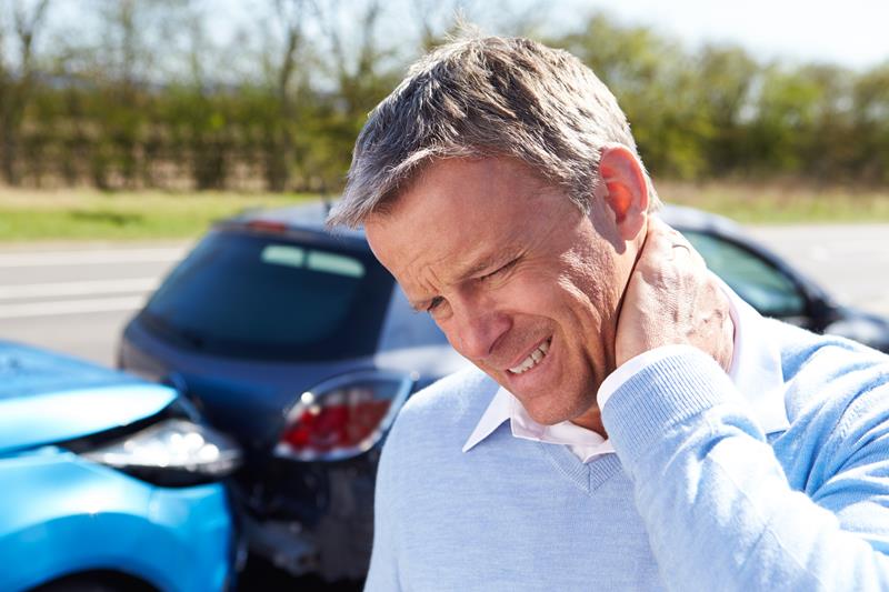 A man with a pained expression clutches his neck with his right hand, standing in front of two cars that appear to have been in a collision.