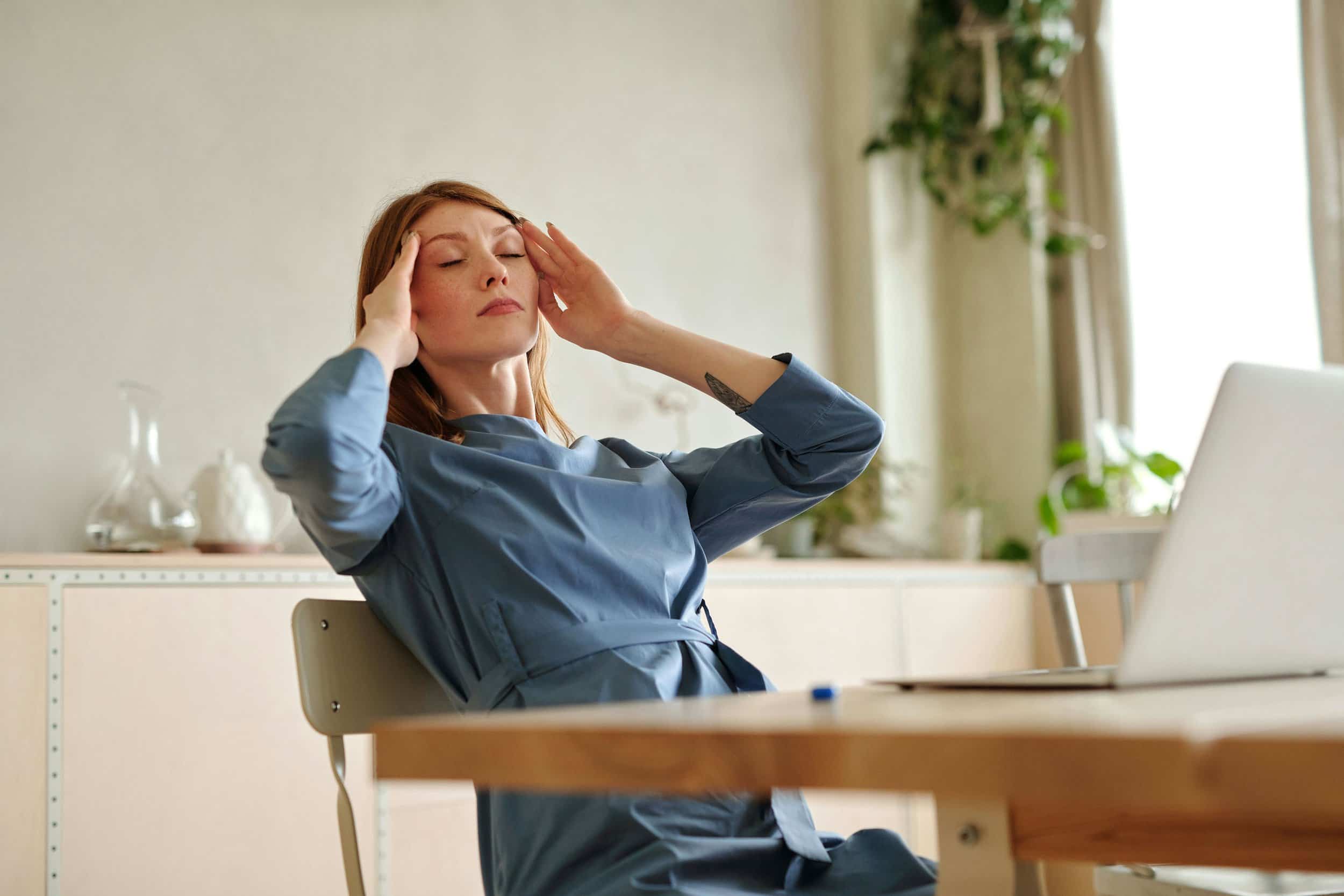 A woman with red hair and freckles, wearing a blue dress, sits with her eyes closed and hands pressed to her temples, indicating a headache or stress. A laptop is on a desk nearby.