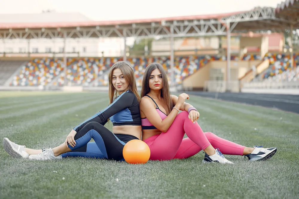 Two women in athletic wear sit back-to-back on a grassy field with an orange ball between them, a stadium in the background.